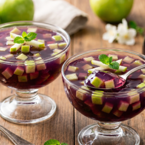 Gelatina de uva de cor roxa profunda em taça elegante, com cubos de maçã verde visíveis no interior e decoração de hortelã, estilo fotografia gastronômica.